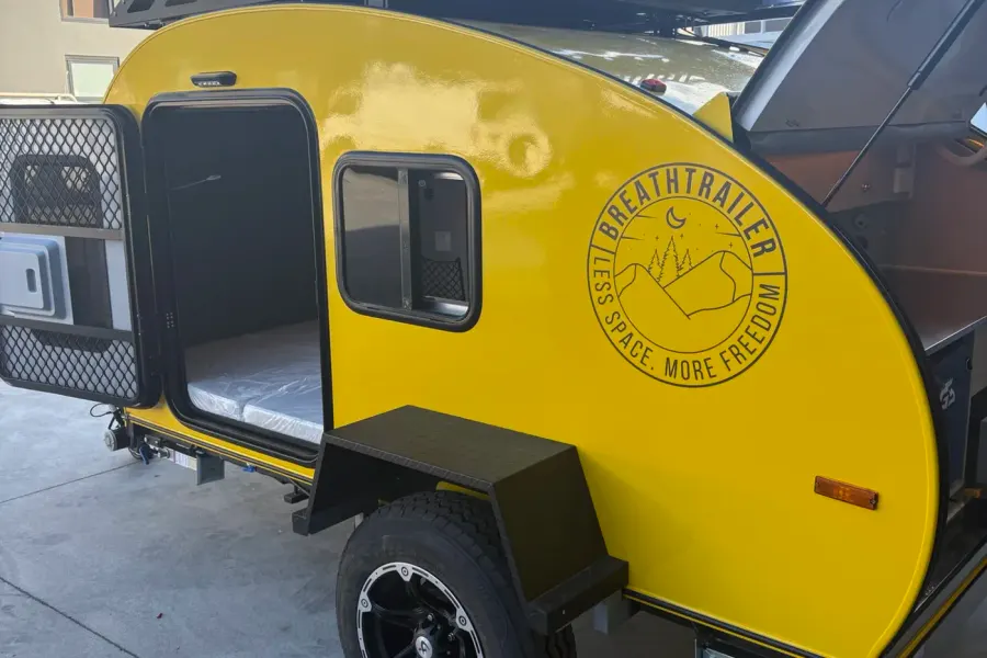 Yellow compact Breath teardrop with side door open showing the mattress, rear hatch raised