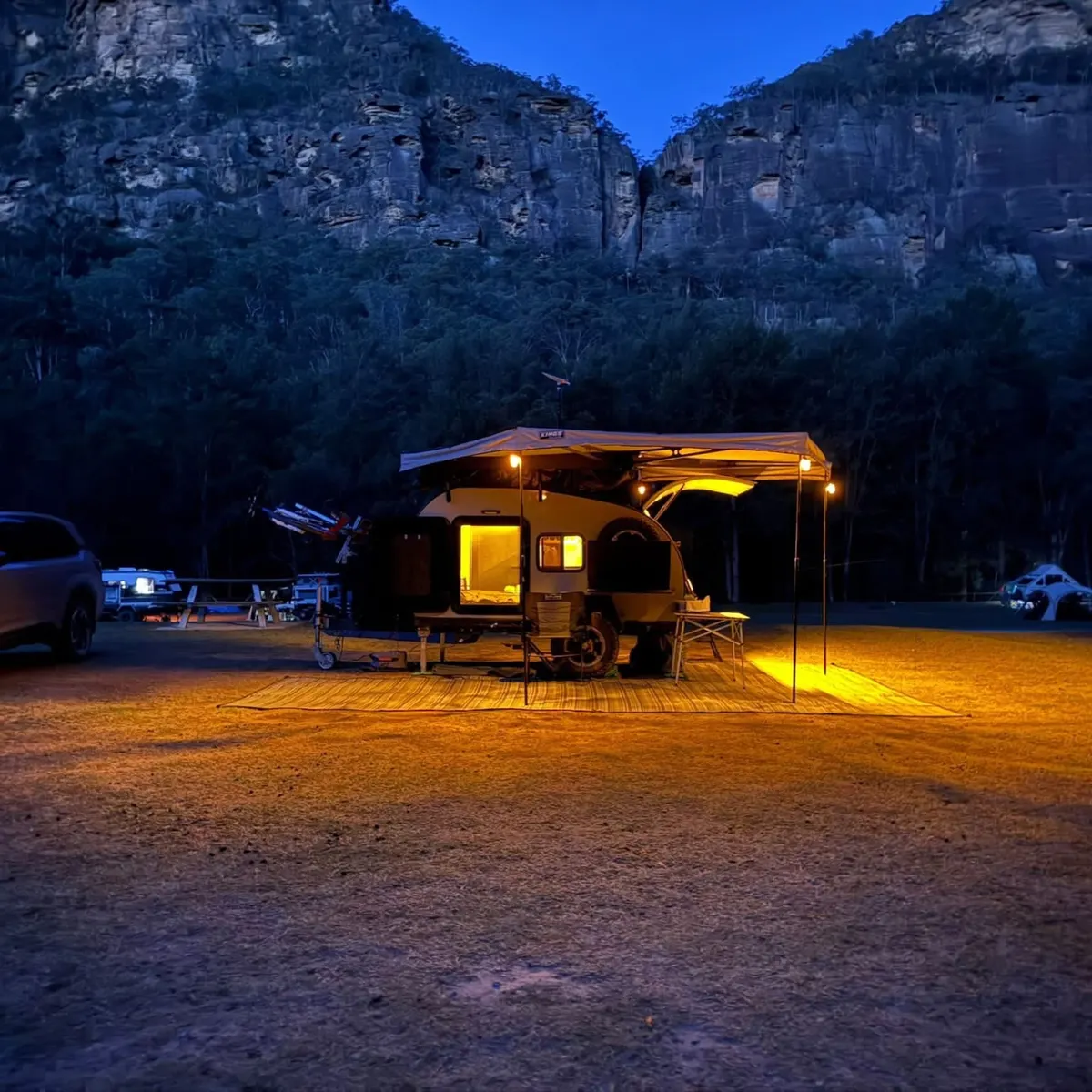 Michael’s Ultra illuminated beneath its awning at a campsite below cliffs at dusk
